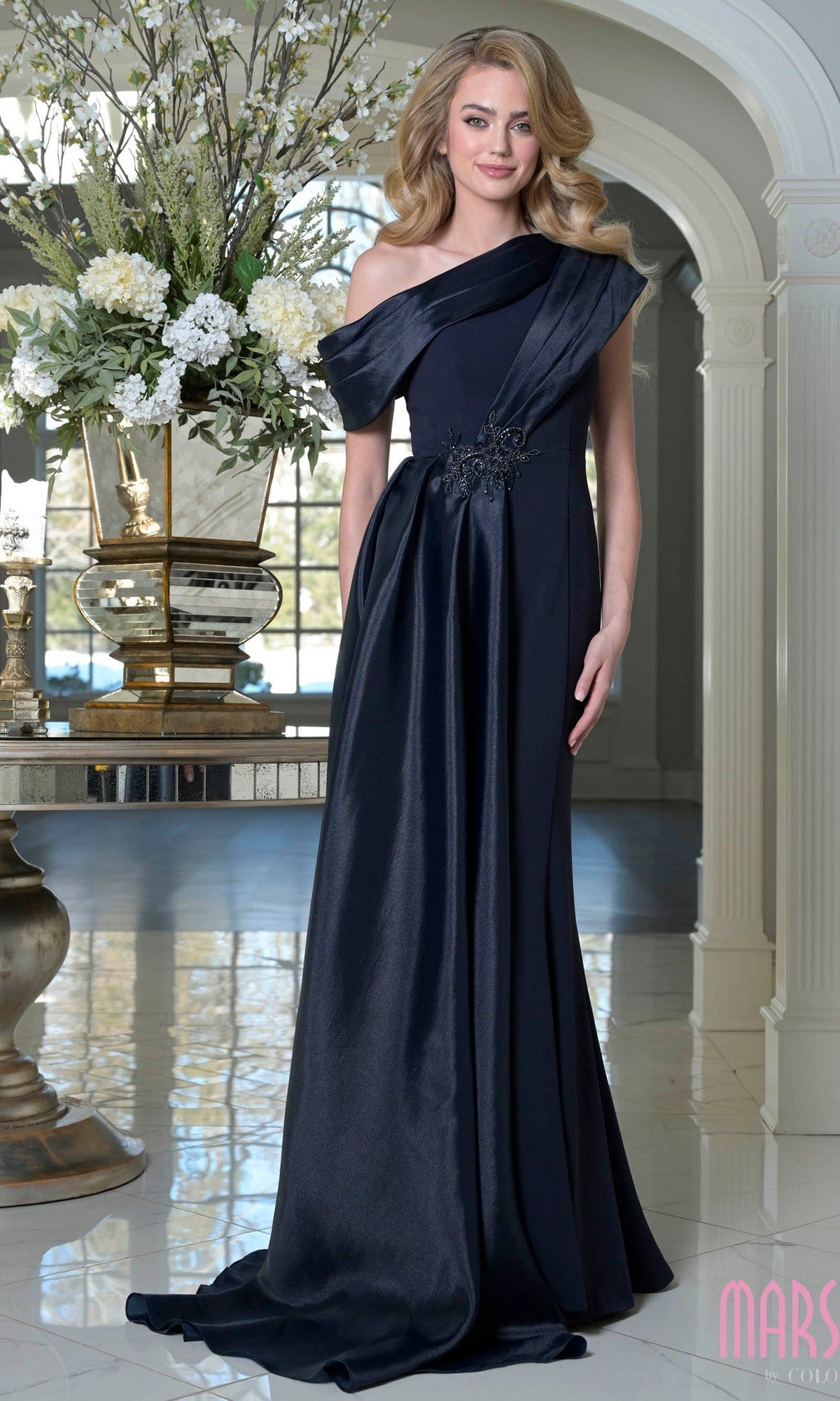 A woman in an asymmetrical navy Marsoni M346 long formal dress stands in an elegant room filled with floral arrangements and decorative vases.